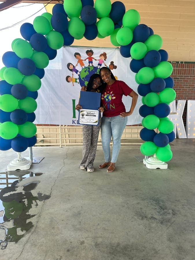 K.I.N. volunteer and a youth holding a certificate under the balloon arch
